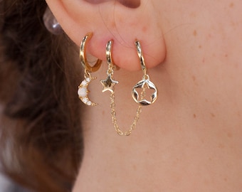 Op de afbeelding: Gouden creolen met hangende bedels, waaronder een halve maan met witte stenen, een ster en een stervormige hanger. De bedels zijn verbonden door een gouden ketting.