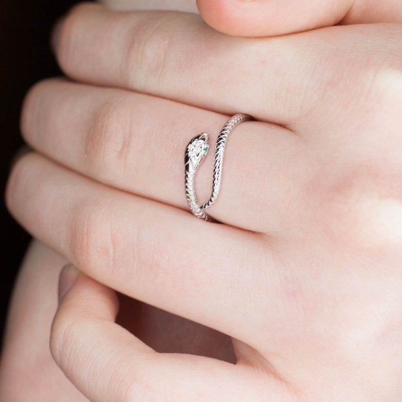 May include: A silver snake ring with a green stone for an eye. The ring is worn on a finger.