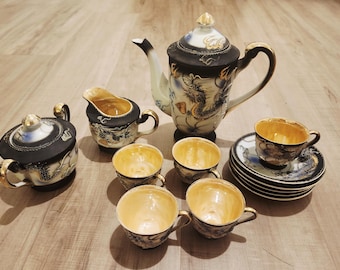 May include: A vintage porcelain tea set featuring a teapot, creamer, sugar bowl, and six teacups with saucers. The set has a black and gold color scheme with dragon and floral designs. The teapot measures approximately 8 inches in height.