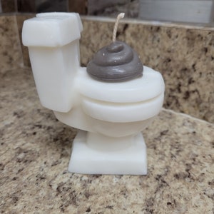 May include: A novelty candle shaped like a white toilet with a gray poop emoji on the seat. The candle has a wick and is sitting on a speckled countertop.