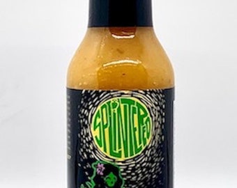 Könnte beinhalten: Eine Glasflasche mit scharfer Sauce mit einem schwarzen Etikett. Das Etikett zeigt eine grüne und gelbe Illustration einer Frau, die eine Ananas hält. Der Text auf dem Etikett lautet "Splintered Luau Pohaku Pineapple - Carolina Reaper - Hot Sauce".