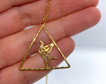 Könnte beinhalten: Eine goldfarbene Halskette mit einem dreieckigen Anhängerrahmen mit offener Mitte. Im Inneren des Dreiecks befindet sich eine stilisierte, geometrische Vogelfigur. Der Anhänger ist an einer zarten, goldfarbenen Gliederkette befestigt.