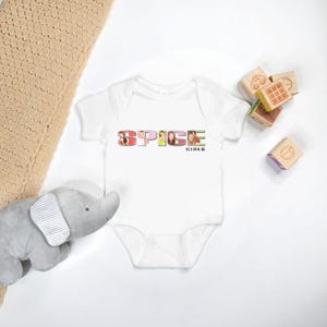 May include: White baby onesie featuring a graphic of the Spice Girls, spelled out with images of each member, and the text "Spice Girls" underneath.