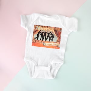 May include: White baby onesie with a printed image of the boy band *NSYNC on the front. The image is from their album *No Strings Attached* and features the band members in a stage setting.