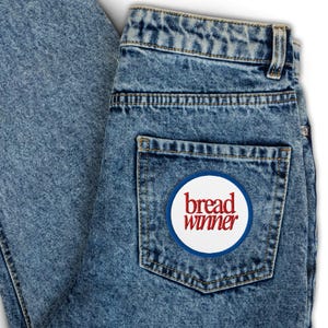 Può includere: Un paio di jeans blu denim con una toppa circolare bianca sulla tasca posteriore. La toppa ha le parole "bread winner" in lettere rosse.