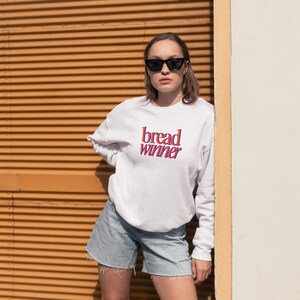 Op de afbeelding: Een wit sweatshirt met de tekst "bread winner" in roze en rode letters. De persoon die het sweatshirt draagt staat voor een muur met oranje horizontale strepen.