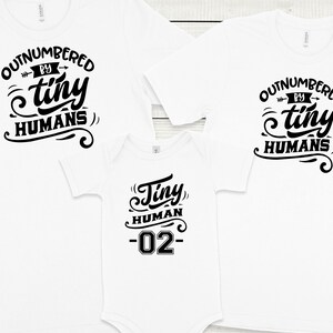 May include: Three white t-shirts with black text. The largest shirt says "Outnumbered by tiny humans". The medium shirt says "Outnumbered by tiny humans". The smallest shirt says "Tiny human -02-"