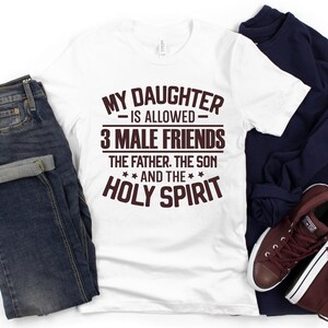 K&ouml;nnte beinhalten: Wei&szlig;es T-Shirt mit dem Text "MY DAUGHTER IS ALLOWED 3 MALE FRIENDS THE FATHER. THE SON AND THE HOLY SPIRIT" in braunen Buchstaben. Das Shirt wird mit blauen Jeans, einem marineblauen Sweatshirt und weinroten Sneakers kombiniert.