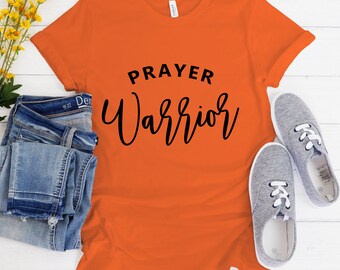 May include: Orange t-shirt with black text that reads "PRAYER WARRIOR". The shirt is laying on a white wooden surface with a pair of gray canvas shoes and blue jeans.