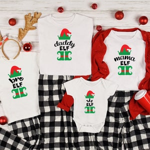 May include: A family of four wearing matching white t-shirts with Christmas elf designs. The shirts say "bro elf", "daddy elf", "mama elf", and "sis elf". The family is wearing black and white plaid pajamas.