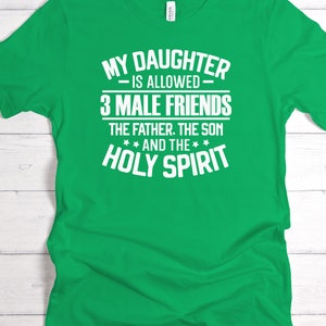 K&ouml;nnte beinhalten: Ein leuchtend gr&uuml;nes T-Shirt mit wei&szlig;em Text. Der Text lautet: "MY DAUGHTER IS ALLOWED 3 MALE FRIENDS THE FATHER. THE SON AND THE HOLY SPIRIT". Das Shirt wird vor einem wei&szlig;en Holzgrund gezeigt.