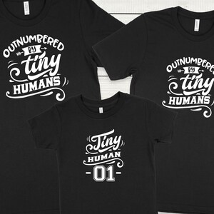 May include: Three black t-shirts with white text. The two larger shirts say "Outnumbered by tiny humans". The smaller shirt says "Tiny human -01-"