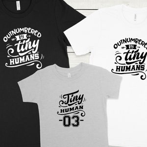 May include: Three t-shirts with a family theme. One black t-shirt says "Outnumbered by tiny humans". One white t-shirt says "Outnumbered by tiny humans". One gray t-shirt says "Tiny human -03".