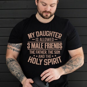 K&ouml;nnte beinhalten: Schwarzes T-Shirt mit dem Text "MY DAUGHTER IS ALLOWED - 3 MALE FRIENDS - THE FATHER, THE SON AND THE HOLY SPIRIT" in Kupferfarbe. Das T-Shirt wird von einer Person mit T&auml;towierungen an den Armen getragen.