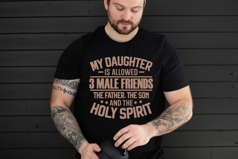K&ouml;nnte beinhalten: Schwarzes T-Shirt mit dem Text "MY DAUGHTER IS ALLOWED - 3 MALE FRIENDS - THE FATHER, THE SON AND THE HOLY SPIRIT" in Kupferfarbe. Das T-Shirt wird von einer Person mit T&auml;towierungen an den Armen getragen.