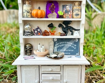 May include: A white miniature hutch with a glass door cabinet, four drawers, and two shelves. The shelves are decorated with a variety of Halloween-themed items, including a pumpkin, a black cat, a dragon, a crystal ball, a Ouija board, and a bottle of potion. The hutch is sitting on a bed of dried grass.