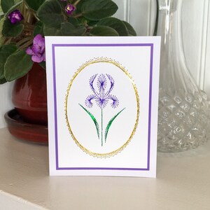 May include: A white card with a purple border and a gold oval frame. Inside the frame is a purple flower with green leaves, embroidered with thread.