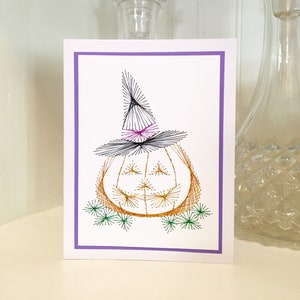 May include: A white card with a purple border features a string art pumpkin wearing a witch's hat. The pumpkin is orange with a black outline and a smiling face. The hat is black with a pink brim and a pointy top.