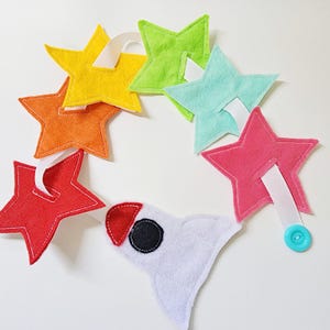 Puede incluir: Una guirnalda de estrellas de fieltro de colores con un cohete de fieltro blanco al final. La guirnalda está enhebrada con una cinta blanca y tiene un botón azul al final.