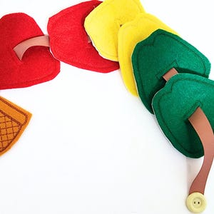 May include: A colorful felt toy featuring a series of connected shapes. The shapes include red, yellow, and green leaf-like forms, along with a brown basket-shaped piece. A brown strap and a button are also visible.