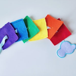 Peut inclure: Un jouet en feutre arc-en-ciel avec un ruban bleu et un bouton. Le jouet est en forme de nuage et comporte une série de carrés de différentes couleurs, notamment rouge, orange, jaune, vert, bleu et violet.