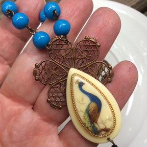 May include: Vintage peacock pendant necklace. The necklace features a teardrop-shaped pendant with a peacock illustration, a bronze-colored filigree flower, and blue bead accents. The pendant is cream-colored with a gold border.
