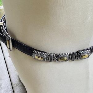 May include: Black belt with silver and gold rectangular details. The belt is made of a textured material and has a silver buckle. The decorative details are evenly spaced along the belt's length, adding a touch of elegance.