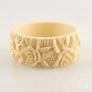 May include: Ivory-coloured bangle bracelet with a floral design. The bracelet features carved roses, leaves, and small bead-like details. The bracelet is approximately 2.5 cm wide.