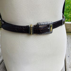 May include: A brown leather belt with a crocodile texture. The belt has a silver and gold-tone buckle with a decorative design. The belt is displayed on a mannequin.