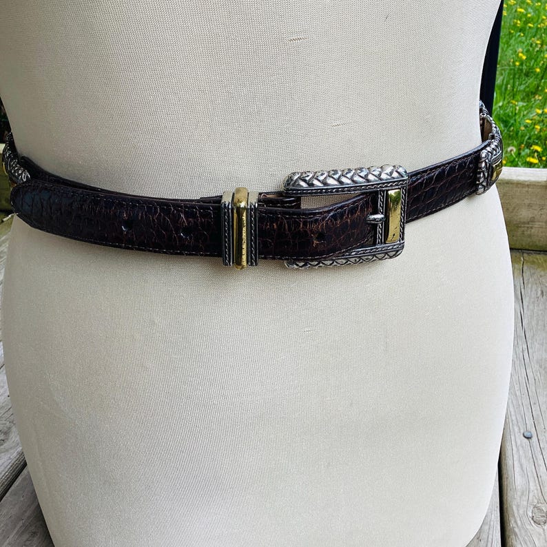 May include: A brown leather belt with a crocodile texture. The belt has a silver and gold-tone buckle with a decorative design. The belt is displayed on a mannequin.
