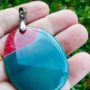 May include: A polished, oval pendant with a silver-coloured bail. The pendant is divided into two sections: a red-pink upper portion and a teal-green lower portion. The stone has a glossy finish and is held in a hand.