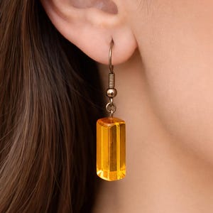 May include: A single gold-toned earring with a faceted, rectangular amber-colored pendant. The earring is suspended from a small hoop and features a coiled spring detail. The earring is shown worn on a person's ear.