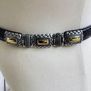 May include: A black belt with silver and gold rectangular accents. The belt has a textured, dark leather strap and features three silver rectangular clasps with gold-coloured centres. The belt is displayed on a mannequin.