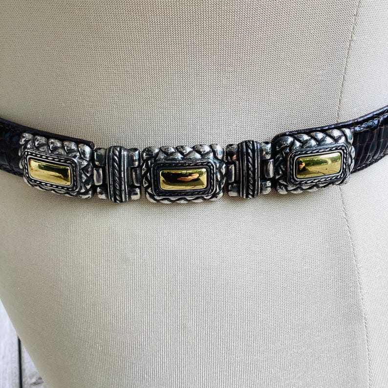 May include: A black belt with silver and gold rectangular accents. The belt has a textured, dark leather strap and features three silver rectangular clasps with gold-coloured centres. The belt is displayed on a mannequin.