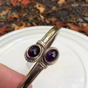 May include: A silver bangle bracelet with two purple amethyst stones set in the centre. The bracelet is open and has a simple design.