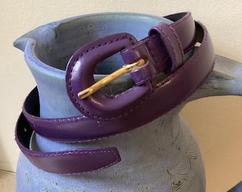 May include: A purple leather belt with a gold buckle. The belt is wrapped around a blue ceramic pitcher.