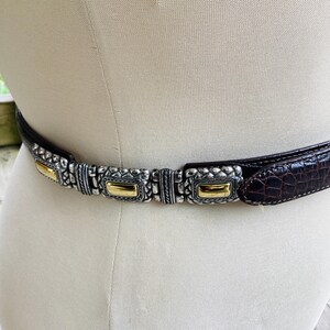 May include: A brown leather belt with a silver and gold-tone metal buckle. The belt has a crocodile texture and is displayed on a mannequin. The buckle features rectangular gold accents and intricate silver detailing.