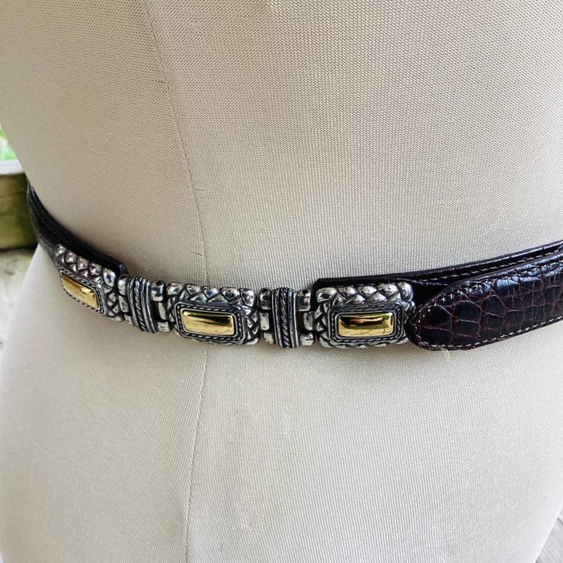 May include: A brown leather belt with a silver and gold-tone metal buckle. The belt has a crocodile texture and is displayed on a mannequin. The buckle features rectangular gold accents and intricate silver detailing.