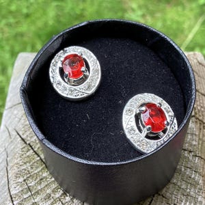 May include: A pair of silver earrings with red oval-shaped stones surrounded by smaller clear stones. The earrings are in a black box.