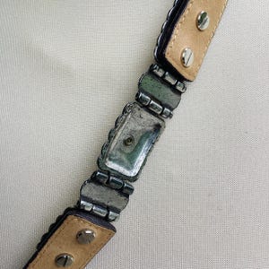 May include: Close-up of a belt with silver-toned metal links and rectangular decorative elements. The belt has a light brown leather backing and is secured with silver screws. The edges of the belt are dark.
