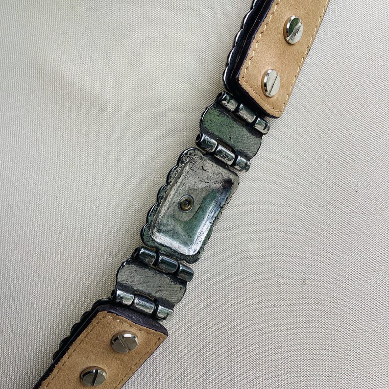 May include: Close-up of a belt with silver-toned metal links and rectangular decorative elements. The belt has a light brown leather backing and is secured with silver screws. The edges of the belt are dark.