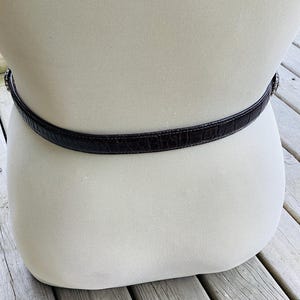 May include: A dark brown belt with a crocodile skin texture is wrapped around a mannequin. The belt has a silver-coloured buckle and is displayed against a neutral background. The belt is a classic accessory.