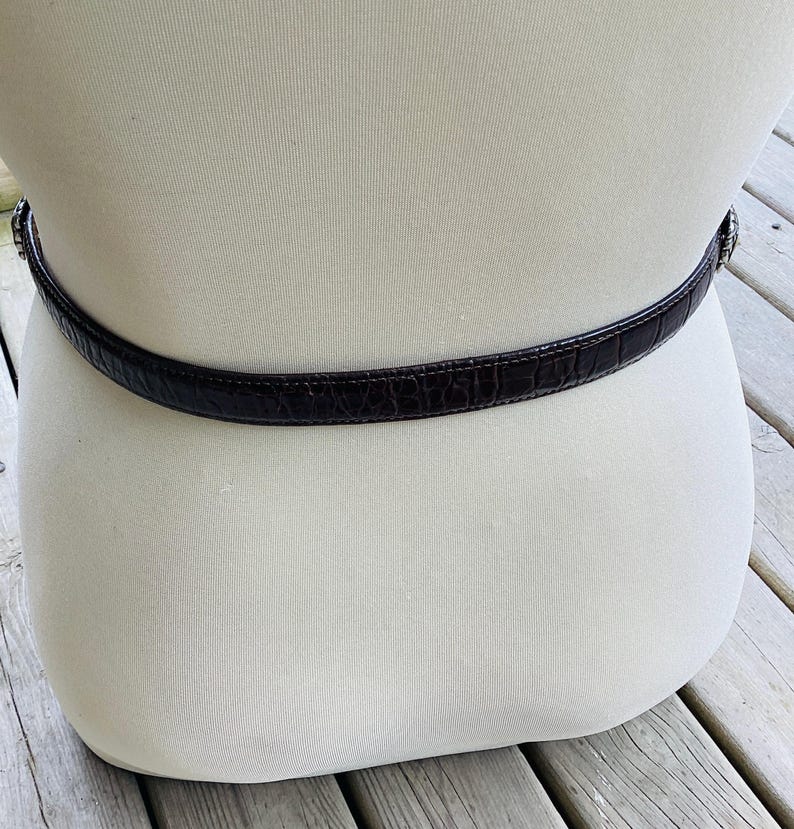 May include: A dark brown belt with a crocodile skin texture is wrapped around a mannequin. The belt has a silver-coloured buckle and is displayed against a neutral background. The belt is a classic accessory.