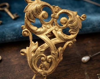 May include: A gold-toned metal hair clip featuring an intricate, openwork design with scrolling leaf and vine motifs. The accessory has a textured, matte finish and a curved, U-shaped base, showcasing a detailed, ornate pattern throughout the piece.