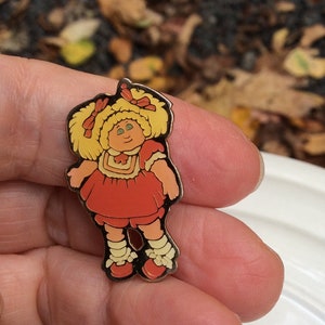 May include: A vintage enamel pin featuring a cartoon character of a girl with blonde hair, wearing a red dress and white socks. The pin is gold-toned and has a detailed design.