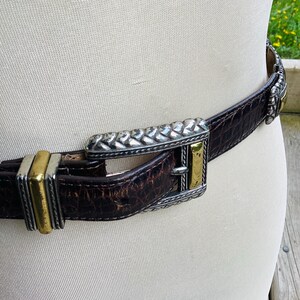 May include: A brown leather belt with a silver and gold buckle. The belt has a textured, crocodile-like pattern. The buckle features a braided silver design and gold accents. The belt is fastened around a mannequin.
