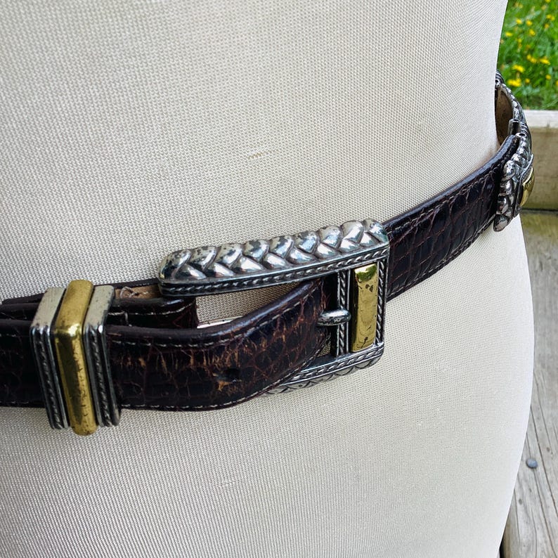 May include: A brown leather belt with a silver and gold buckle. The belt has a textured, crocodile-like pattern. The buckle features a braided silver design and gold accents. The belt is fastened around a mannequin.