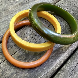 May include: Three circular bangle bracelets stacked together. One bracelet is a deep olive green, one is a bright mustard yellow, and the third is a warm burnt orange. Each bangle has a smooth, polished, and rounded surface with a glossy finish.