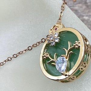 May include: A gold necklace with a green jade pendant featuring a floral design with clear and red crystals.