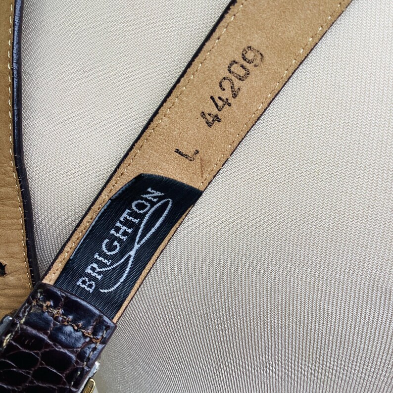 May include: Close-up of a brown leather belt with a textured, crocodile-like pattern. The belt has a black tag with the brand name "BRIGHTON" in white script. The size is "L" and the style number is "44209".